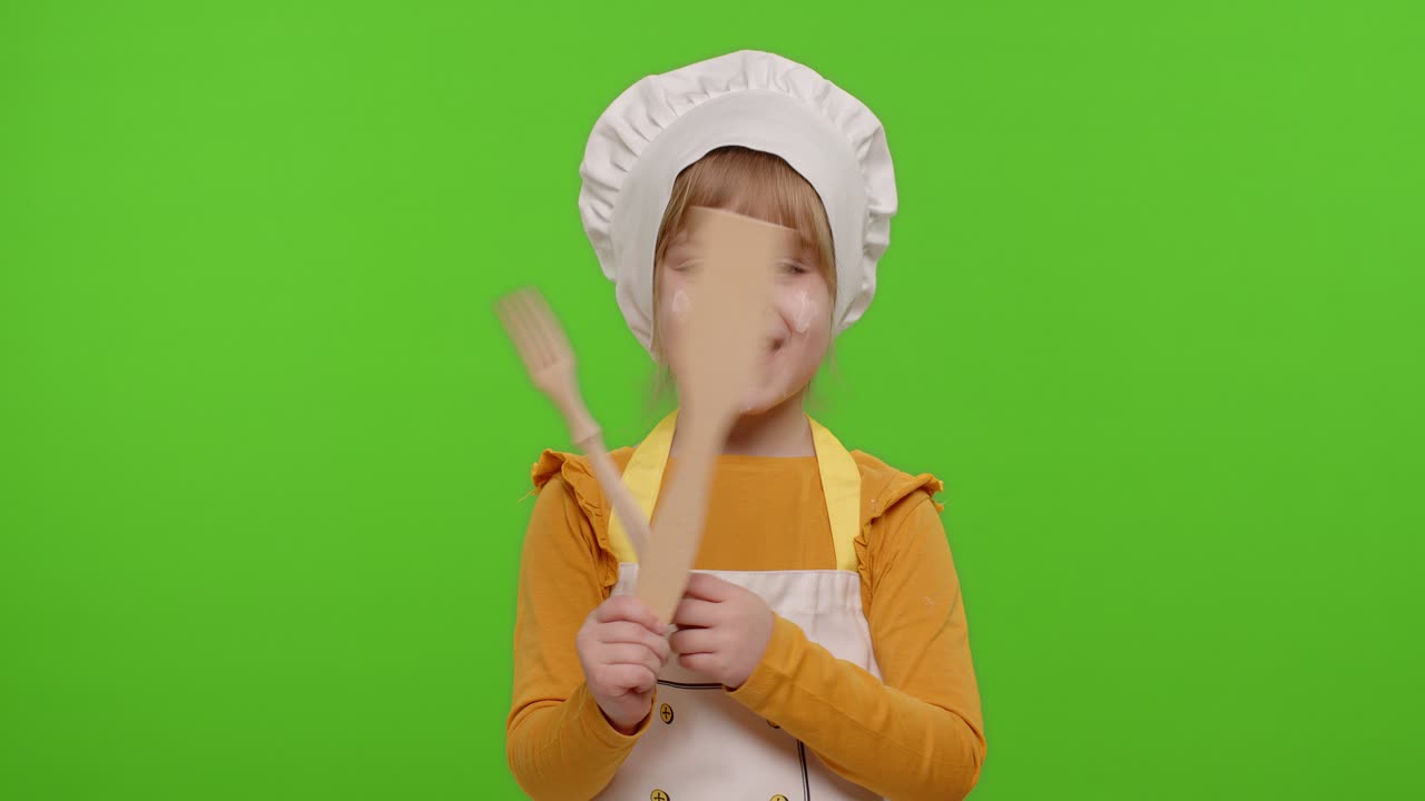 Bambina vestita da cuoca che mostra una forchetta di legno e una spatola, sorridente, che balla su chroma key.