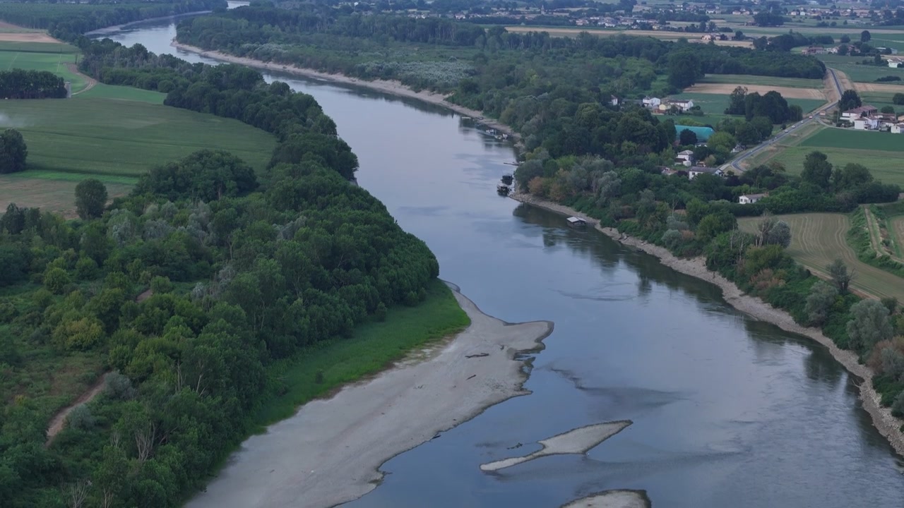 Il fiume Po mostra livelli d'acqua ridotti e sponde esposte vicino alla diga di Isola Serafini a Monticelli d'Ongina, Piacenza, con campi agricoli e rive boscose, inquadratura verso il basso che rivela lo skyline di Cremona