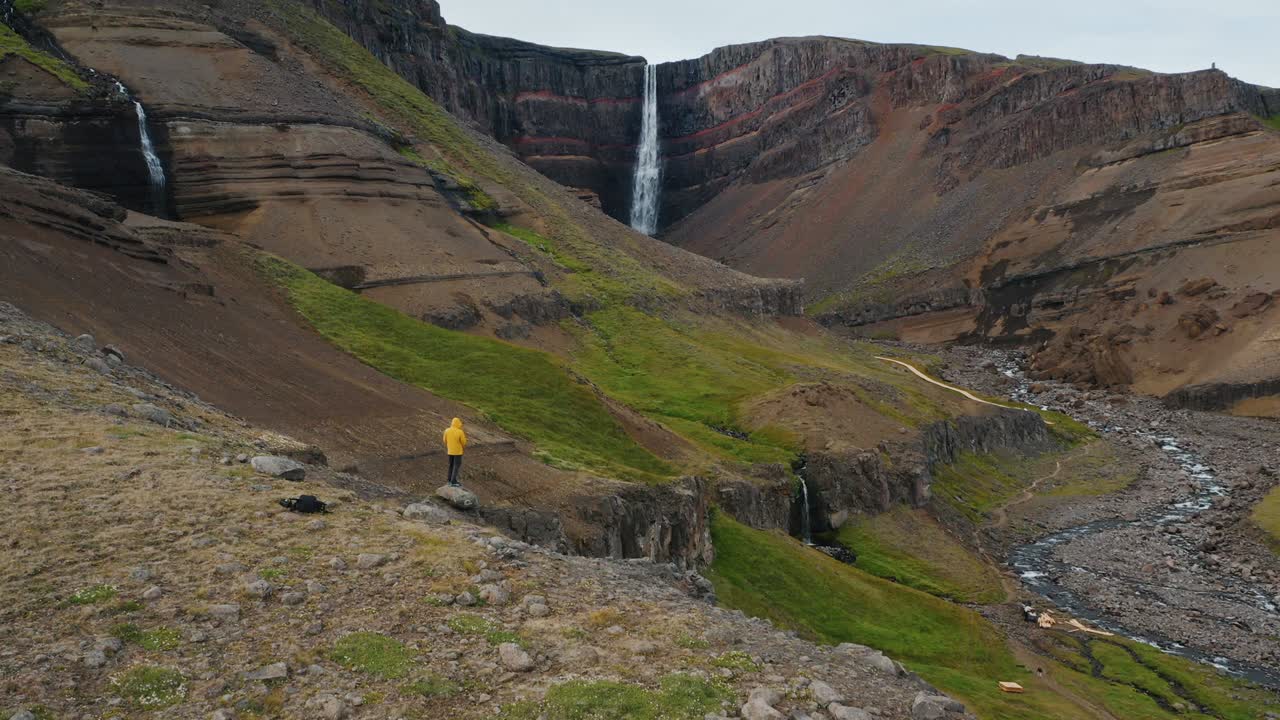 catarata de hengifoss, islandia. vuelo aéreo por encima del hombre turista en chaqueta amarilla en la colina disfrutando del cañón y el río de la montaña