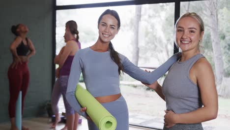 Diverse-female-friends-talking-and-smiling-at-camera-while-standing-in-yoga-studio