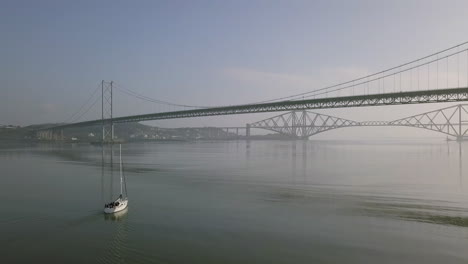 Aerial-footage-of-a-sailing-boat-travelling-beneath-the-old-Forth-Road-Bridge-at-South-Queensferry-on-a-sunny-day-in-West-Lothian,-Scotland