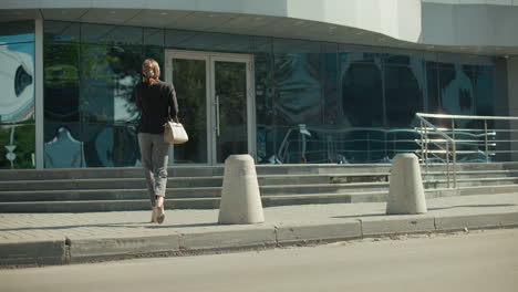 back view of woman holding handbag walking to work while on phone call, approaching modern building with reflective glass panels showing parked cars and metal railing