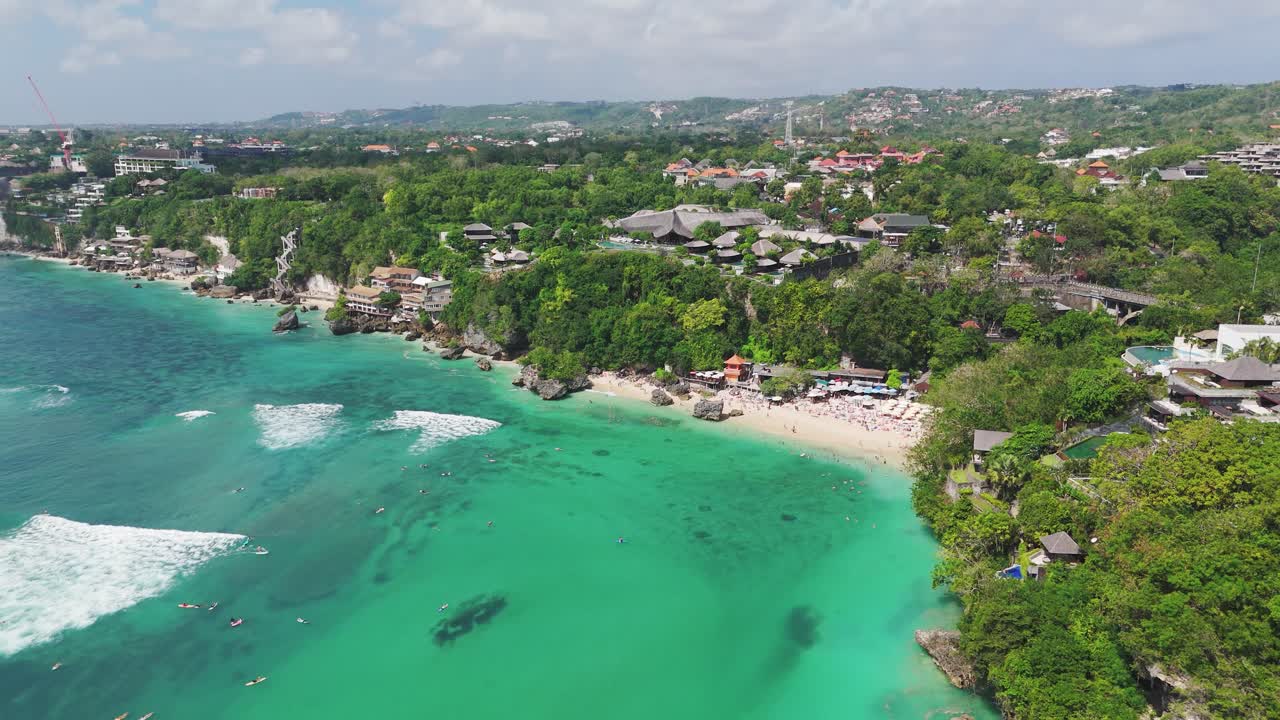 Surfers paddle in turquoise water below cliffs at Padang Padang Beach, Uluwatu, drone pullback