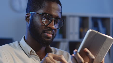 close up view of businessman in glasses watching something on the tablet and taping on hte screen in the office at night