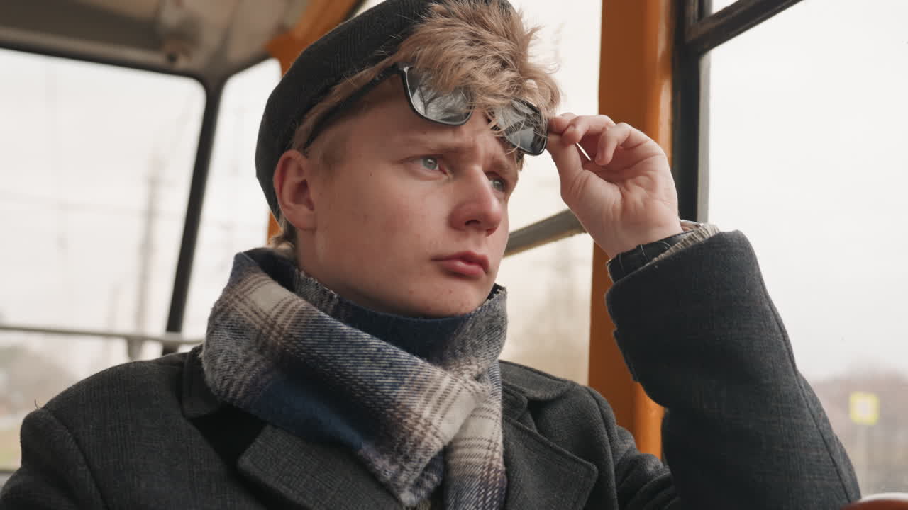 Weißer Kreativer, der auf der Straßenbahn die Sonnenbrille zurechtrückt, die Stadt durch ein regennasses Fenster scannt, Strickschal und Mütze trägt, selbstbewusster Fokuswechsel von stiller Reflexion zur Inhaltssuche, filmisches Licht und gedämpfte Farben
