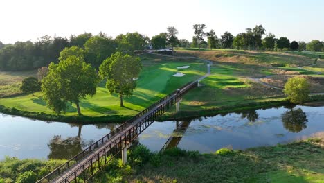 sunlit golf course in the usa, featuring a wooden bridge over a serene river, adjacent green fairways, and mature trees