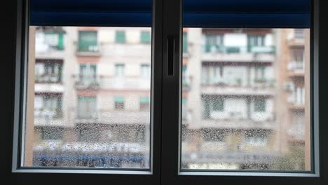 view from inside the room of wet window surface after heavy thunderstorm, closed window with blue shutters on background of blurred residential building in rainy weather day. sad mood during heavy