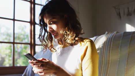 Happy-mixed-race-woman-sitting-on-couch-using-smartphone-in-sunny-living-room