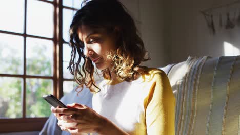 Glückliche-Frau-Mit-Gemischter-Abstammung,-Die-Auf-Der-Couch-Sitzt-Und-Ihr-Smartphone-Im-Sonnigen-Wohnzimmer-Nutzt