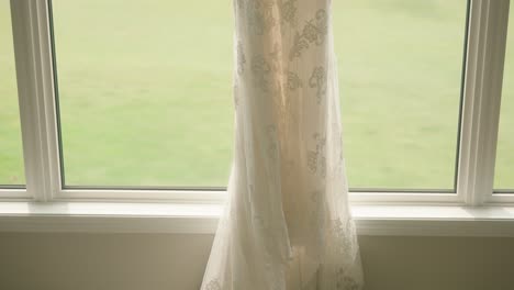 white floral wedding dress hangs in a window with a lakeside view and flower bouquets