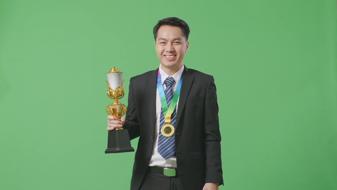 슈트와 타이를 입은 아시아 사업가, 황금 메달을 입고 손에 황금 트로피를 들고, 스튜디오의 녹색 화면 배경에서 카메라에 미소 짓는 첫 번째 우승자입니다.
