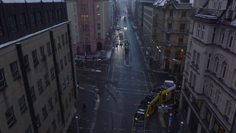 Ascending-aerial-footage-of-tram-slowly-passing-through-crossroad,-turning-right.-Winter-city-at-dusk.-Berlin,-Germany