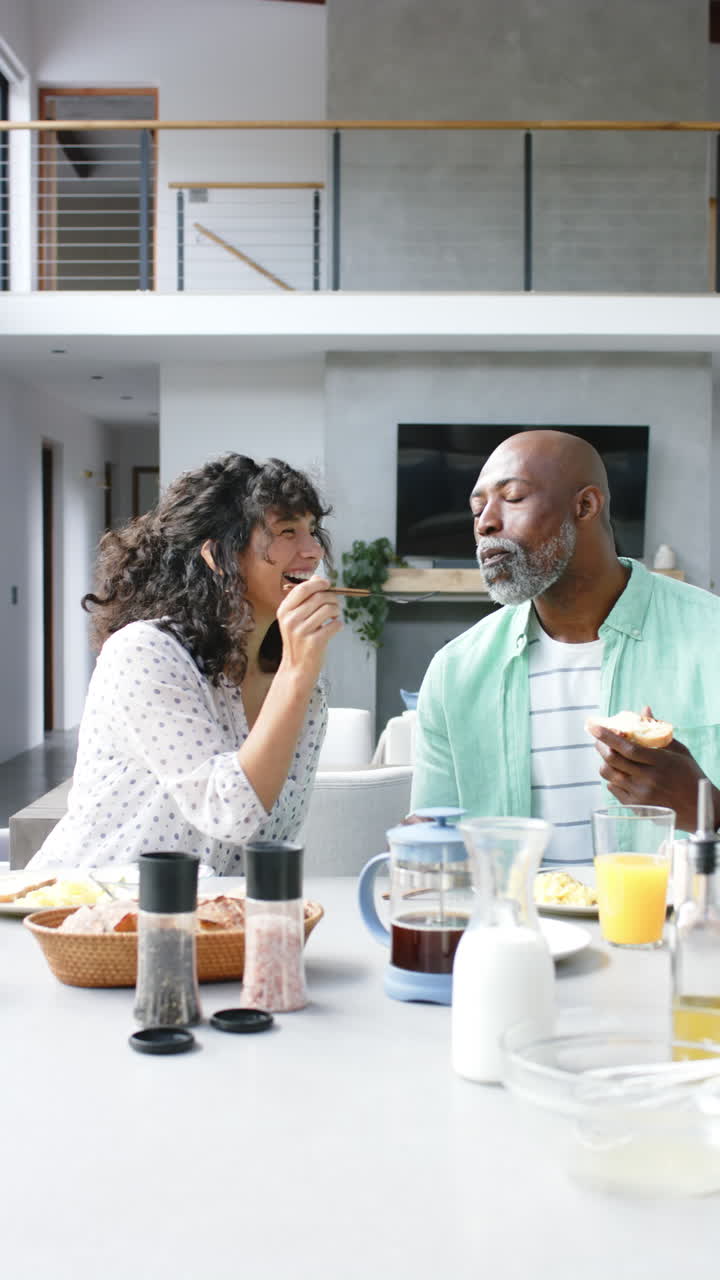 Vertical video of happy diverse mature couple eating breakfast together at home, slow motion