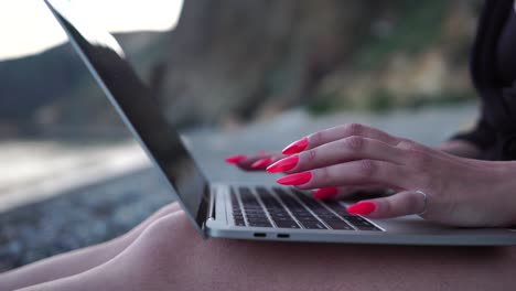 mujer de negocios exitosa con manicura roja brillante escribiendo en el teclado de la computadora portátil al aire libre en la playa con vista al mar. cerca de las manos de la mujer escribiendo en la computadora. freelance, nómada digital, viajes y vacaciones