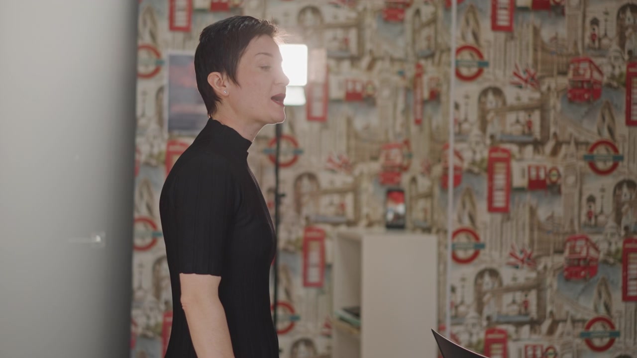 Insegnante di canto che conduce il riscaldamento in studio; l'istruttore dai capelli corti si trova in una sala prove a fantasia, guidando la respirazione e la postura, con espressione concentrata e retroilluminazione soffusa, un momento di insegnamento intimo.