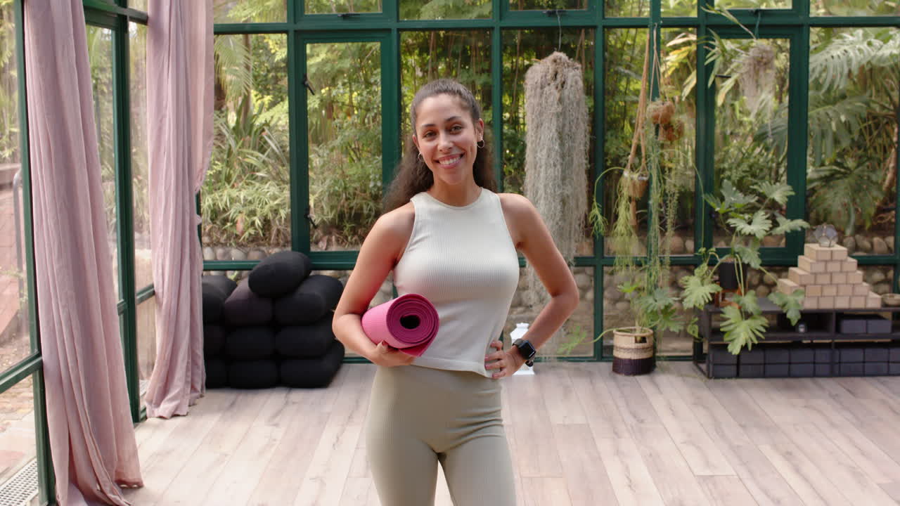 Holding yoga mat, woman smiling and posing in glass house yoga studio