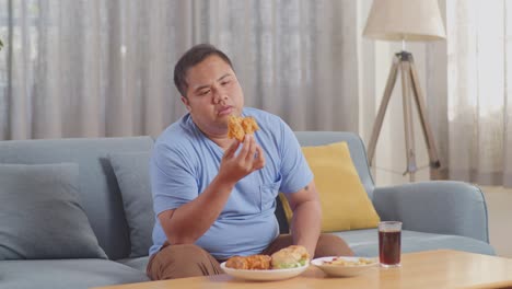 a fat asian man shaking his head being bored having fried chicken while eating fast food on a sofa in the living room at home