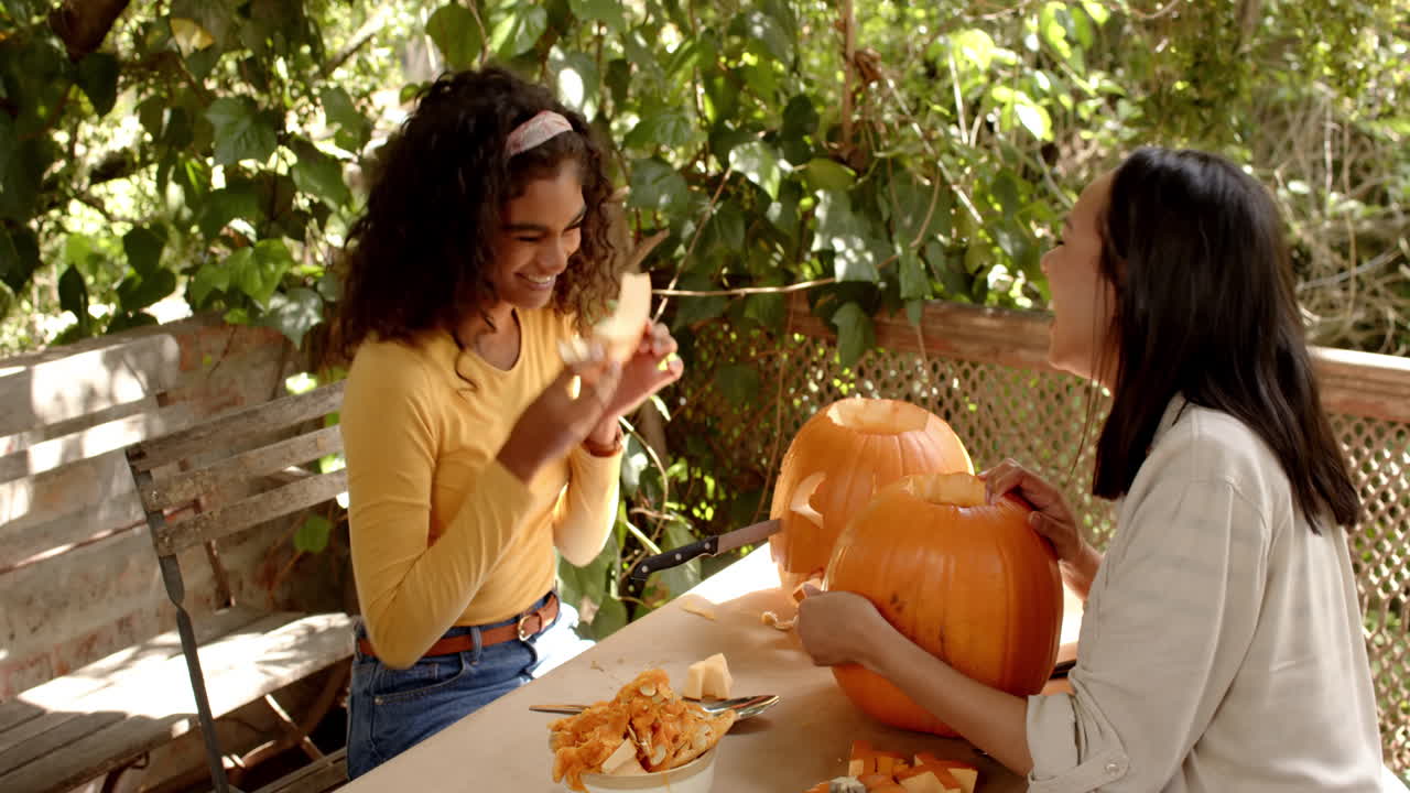 Halloween time, two multiracial female friends carving pumpkins on a porch