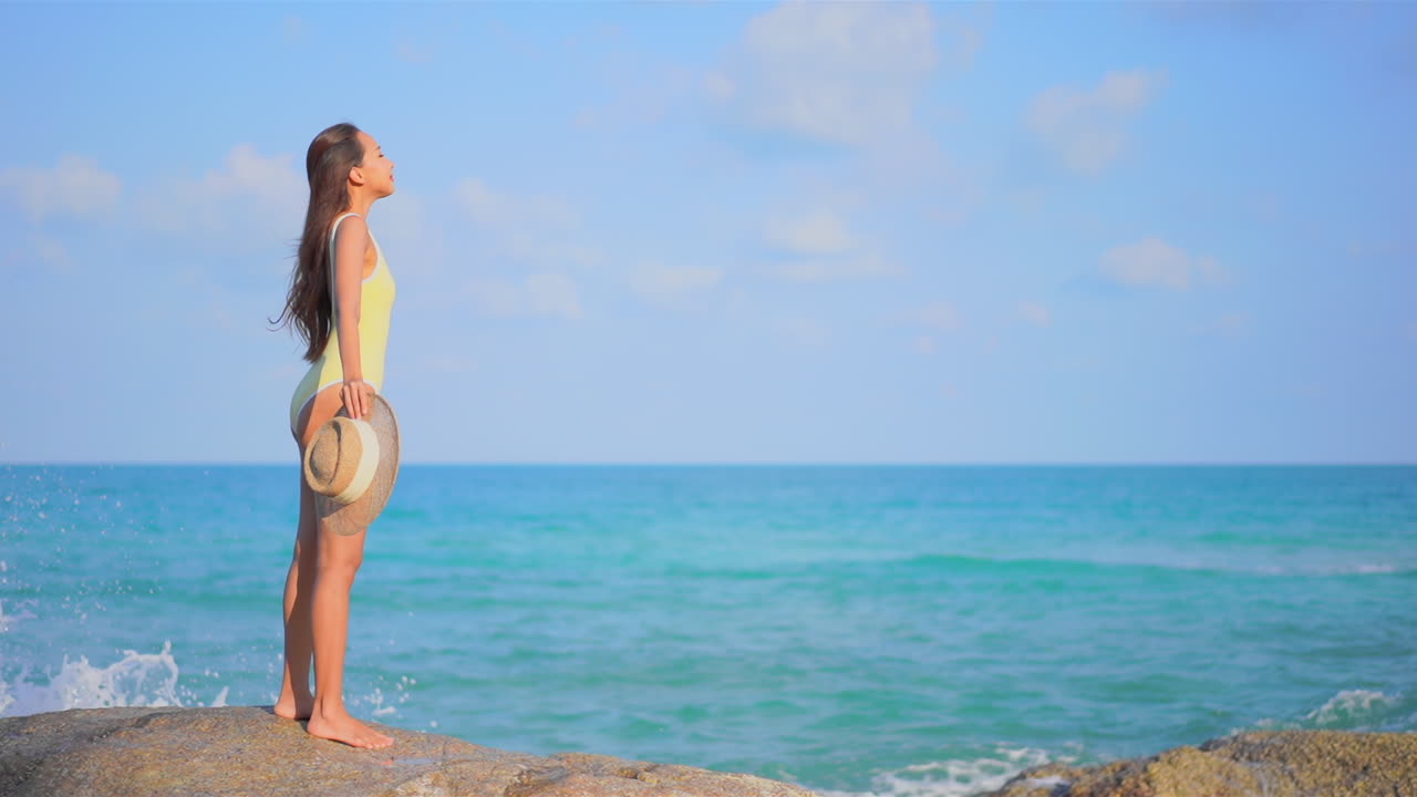 들어오는 파도가 내려다보이는 거대한 바위 위에 서서 노란색 수영복을 입은 젊은 여성이 바닷바람을 즐긴다
