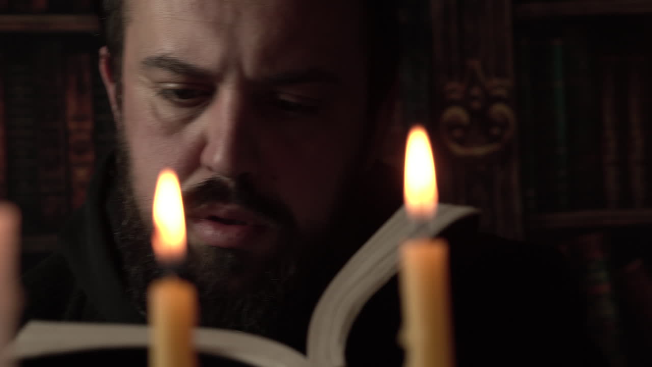 Uomo con barba che legge un libro in biblioteca alla luce di una candela, primo piano