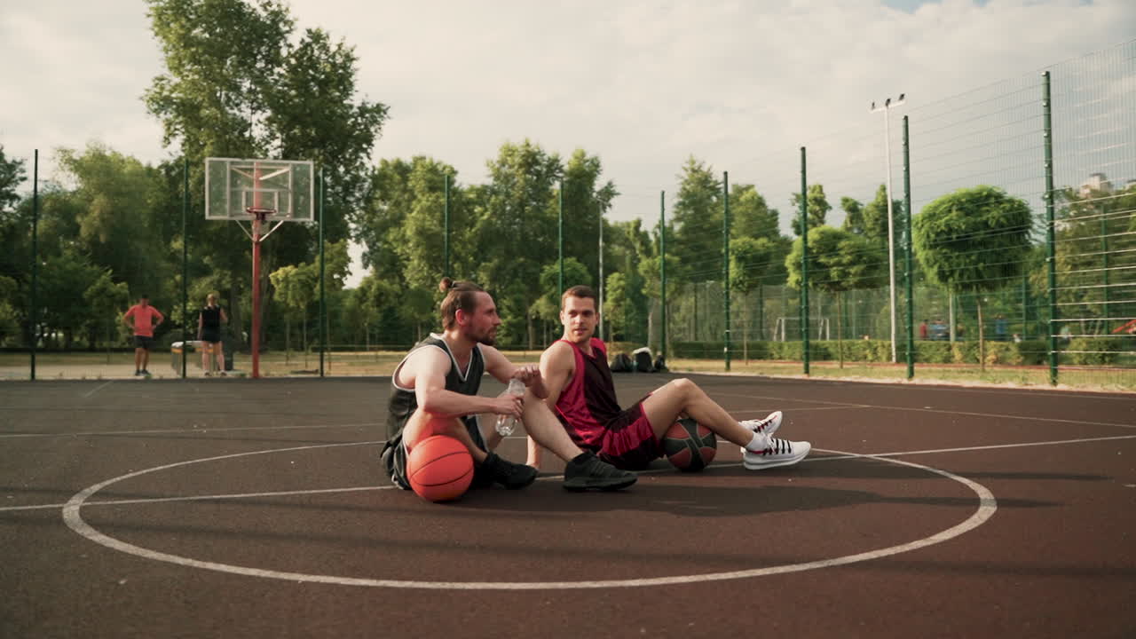 dos jugadores de baloncesto sentados en el centro de una cancha de baloncesto al aire libre, tomando un descanso, bebiendo agua y hablando entre ellos