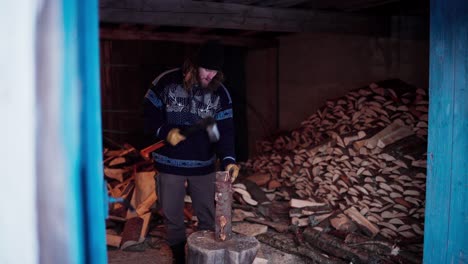 man chopping wood with axe in a barn with pile of firewood