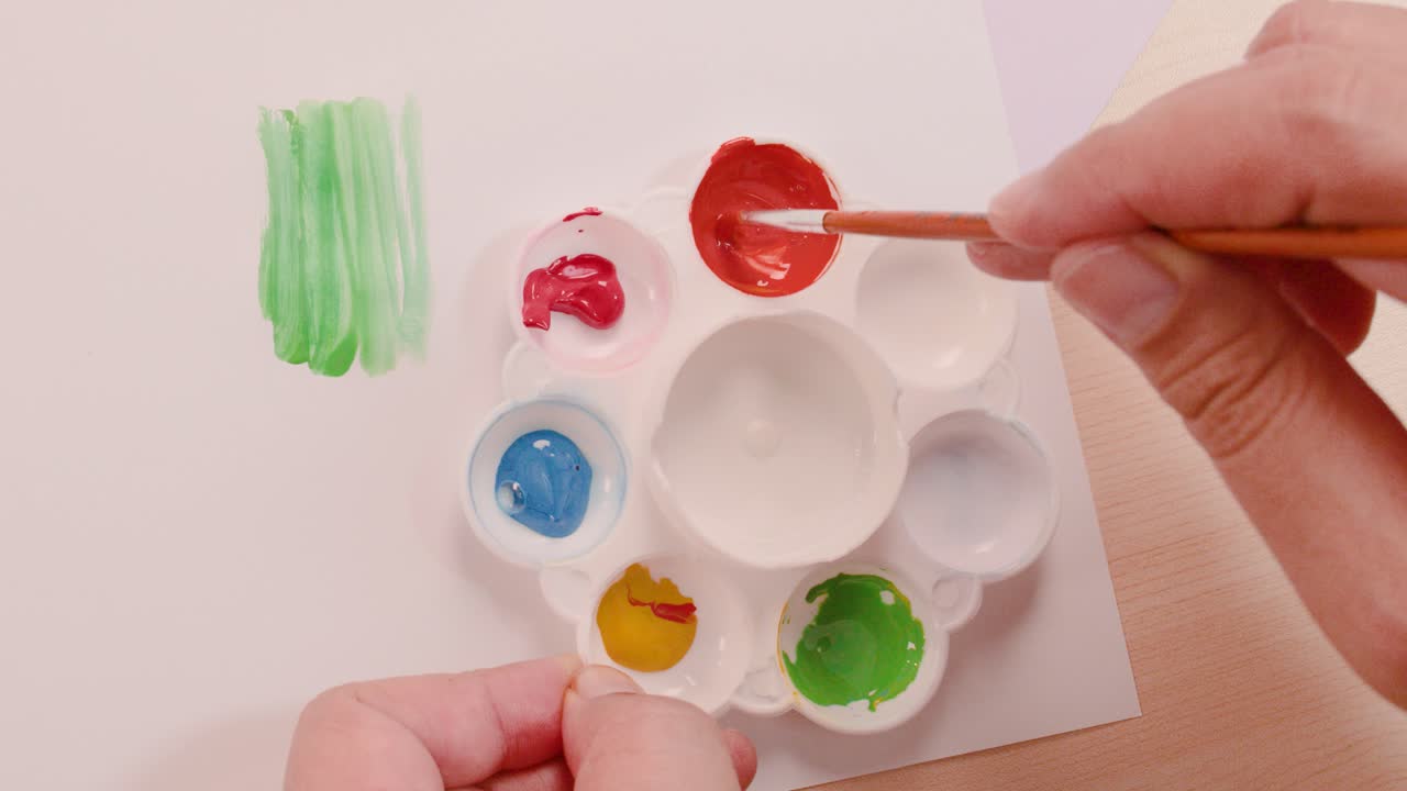 Overhead view of hand mixing orange paint in palette with brush, bright even lighting