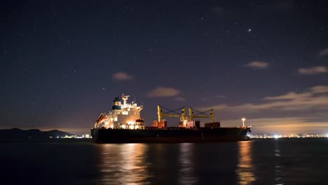 a large cargo vessel is illuminated by lights as it sits in the water during nighttime. the bright stars above contrast with the bustling harbor, showcasing activity along the coastline.
