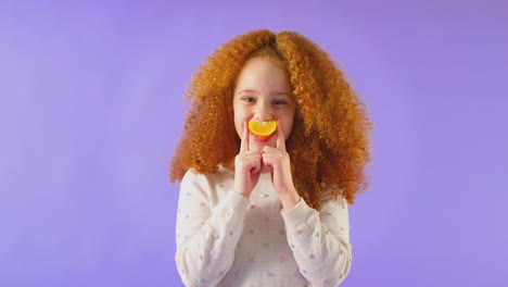 Retrato-De-Estudio-De-Una-Chica-Sosteniendo-Naranja-Para-La-Boca-Sonriente-Contra-El-Fondo-Púrpura