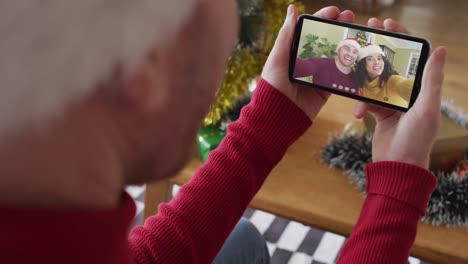 Kaukasischer-Mann-Mit-Weihnachtsmütze-Nutzt-Smartphone-Für-Weihnachtsvideoanruf,-Mit-Paar-Auf-Dem-Bildschirm