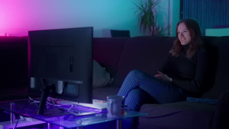 Young-white-woman-laughing-while-watching-TV-at-home-at-night-in-living-room-illuminated-with-purple,-pink-and-teal-colors