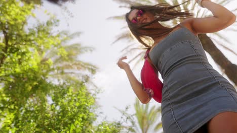 young woman in denim dress and sunglasses dancing under palm trees