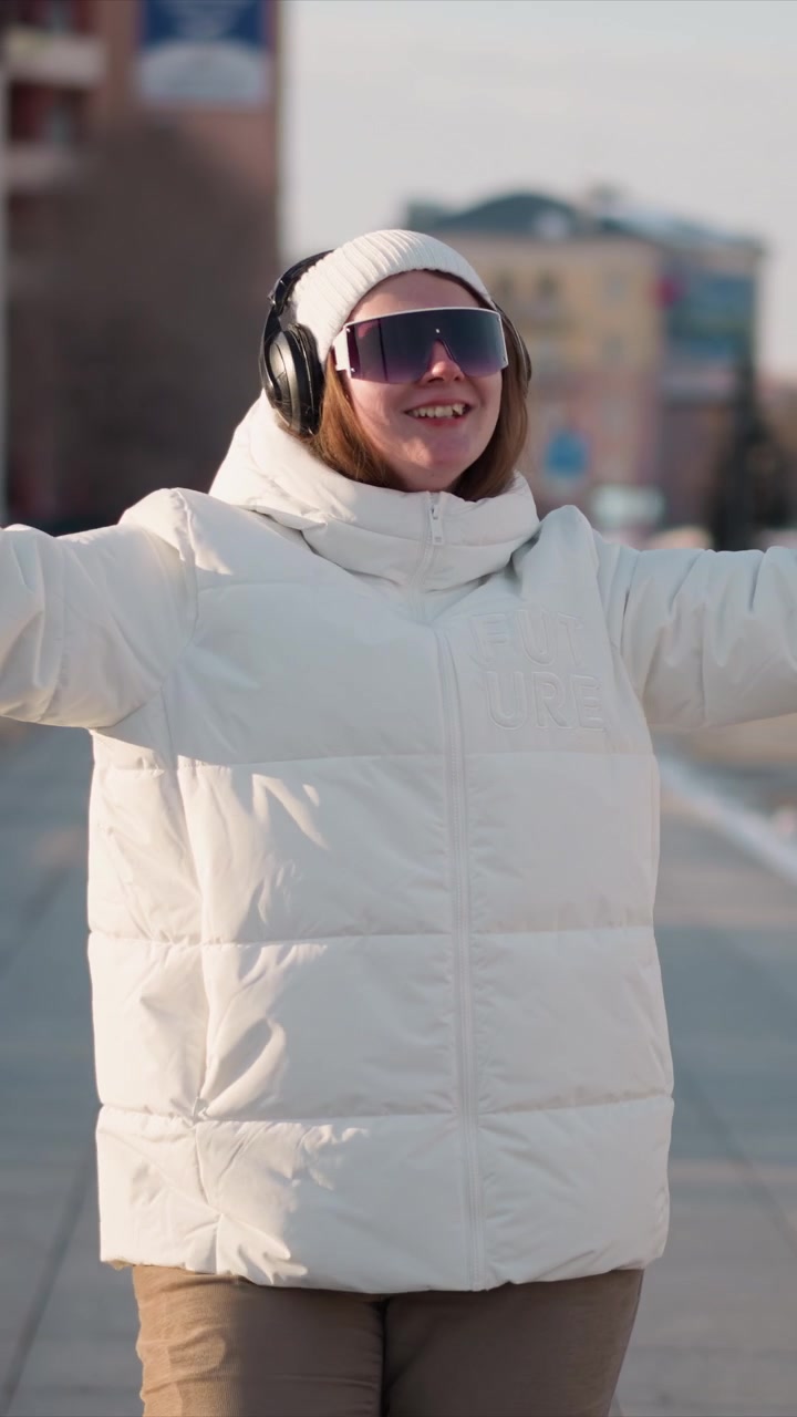 Woman wearing white puffer jacket and headphones dances energetically on urban sidewalk during daylight. Her movements are lively with arms raised or extended as woman performs dance