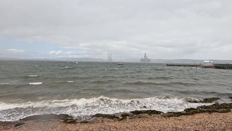 wind-driven waves break on a rocky beach under overcast skies, with sailboats and fishing vessels visible offshore near cromarty, scotland. static wide shot, natural light