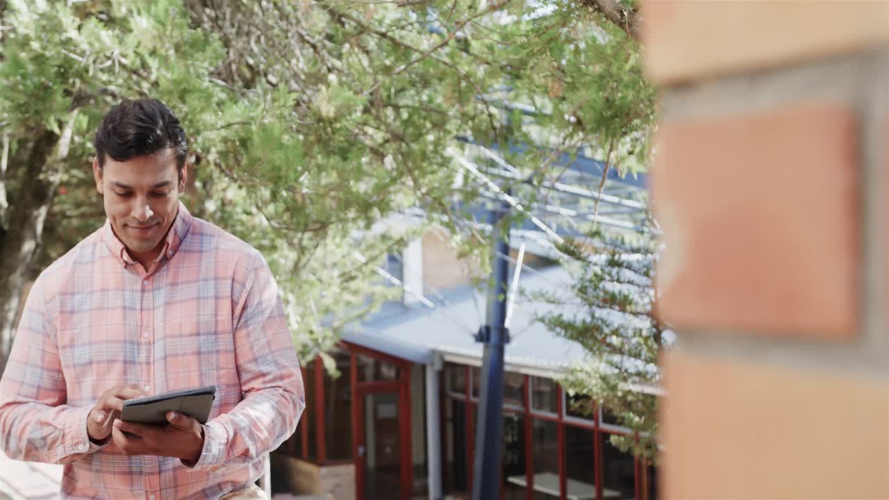 retrato de un feliz maestro de sexo masculino biracial usando una tableta digital de pie cerca de una escuela primaria