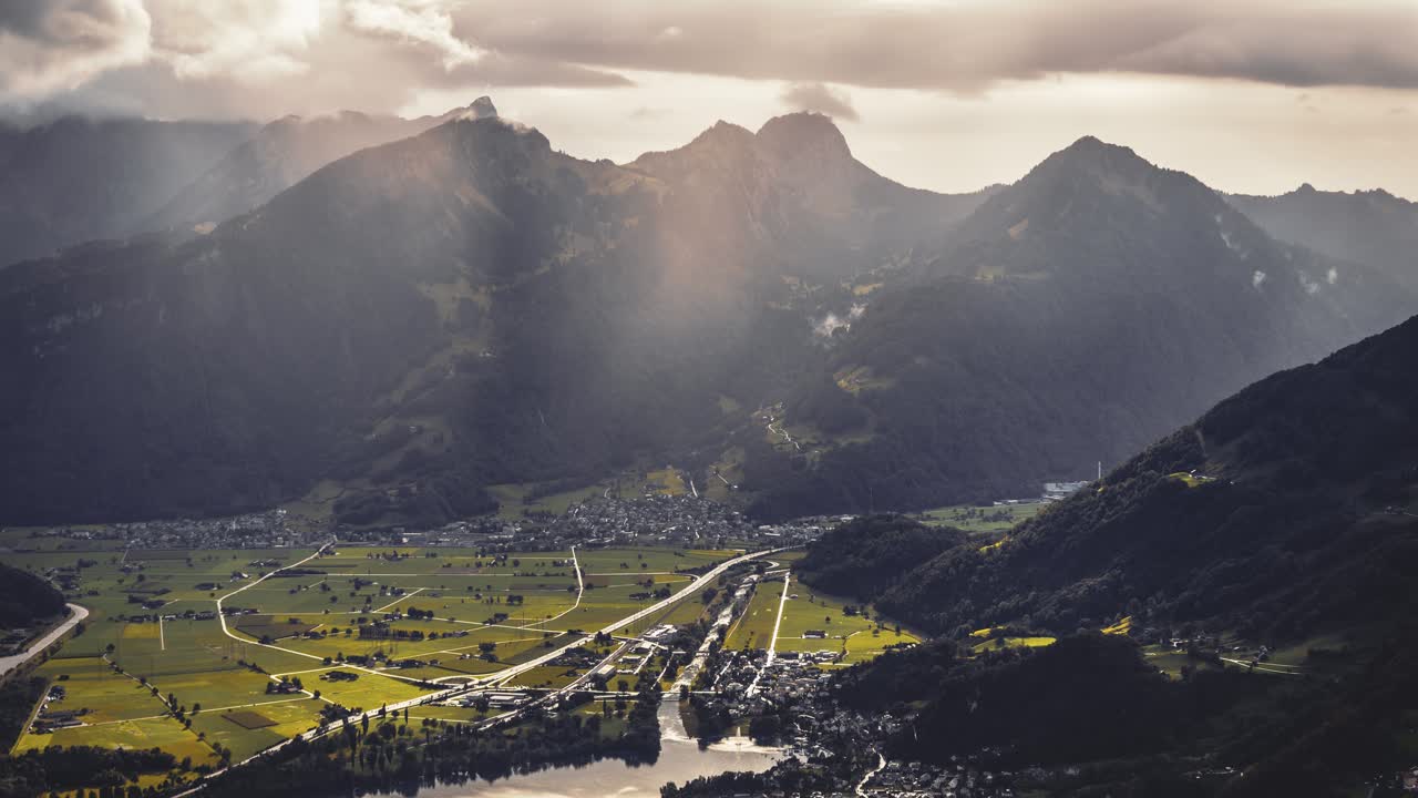 Sunbeams break through rain clouds, casting a golden glow near the town of Amden (St. Gallen, Switzerland). A small town is located on the lakeside with lush green valley around it (Timelapse).