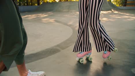un tipo moreno con una camiseta gris ayuda a una chica con una camiseta púrpura y pantalones a rayas a montar patines rosados de 4 ruedas en un parque de patinaje en un piso de hormigón en verano