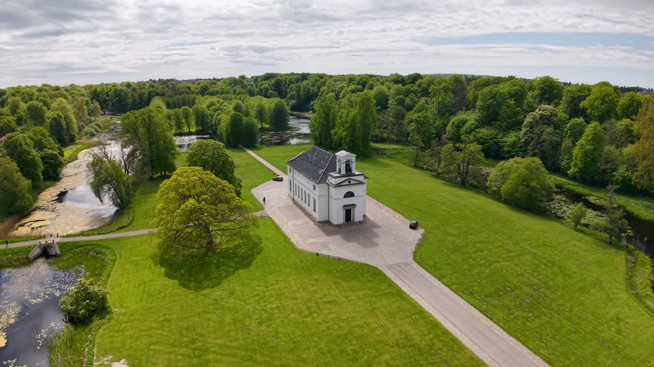 erial drone view of Hørsholm Church with surrounding greenery, reflective ponds, and a bright open sky creating a peaceful landscape