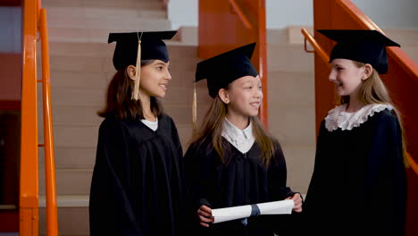 three happy preschool female students in cap and gown standing on stairs, holding diploma and talking together at the graduation ceremony 1