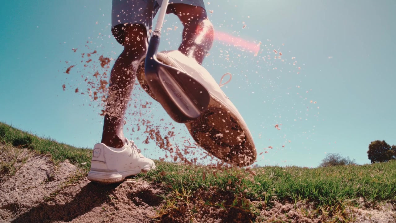 Golfista colpisce il wedge sulle scarpe per pulire i tacchetti uscendo da un bunker di sabbia del deserto, mentre i granelli si disperdono nella luce, filmato dal basso e da vicino al rallentatore, in 4k a 800fps in una giornata di sole.