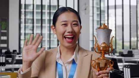 Close Up Of Asian Business Woman In A Suit With A Gold Medal And Trophy Waving Her Hand And Smiling To Camera As The First Winner In The Office