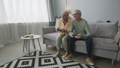 happy middle aged arabic woman and senior man playing video game while sitting on sofa at home