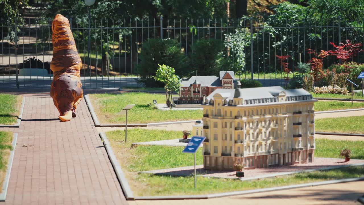 Person in funny dragon costume outdoors. Back view of dinosaur walking in the summer park along toyful buildings in sunny day.