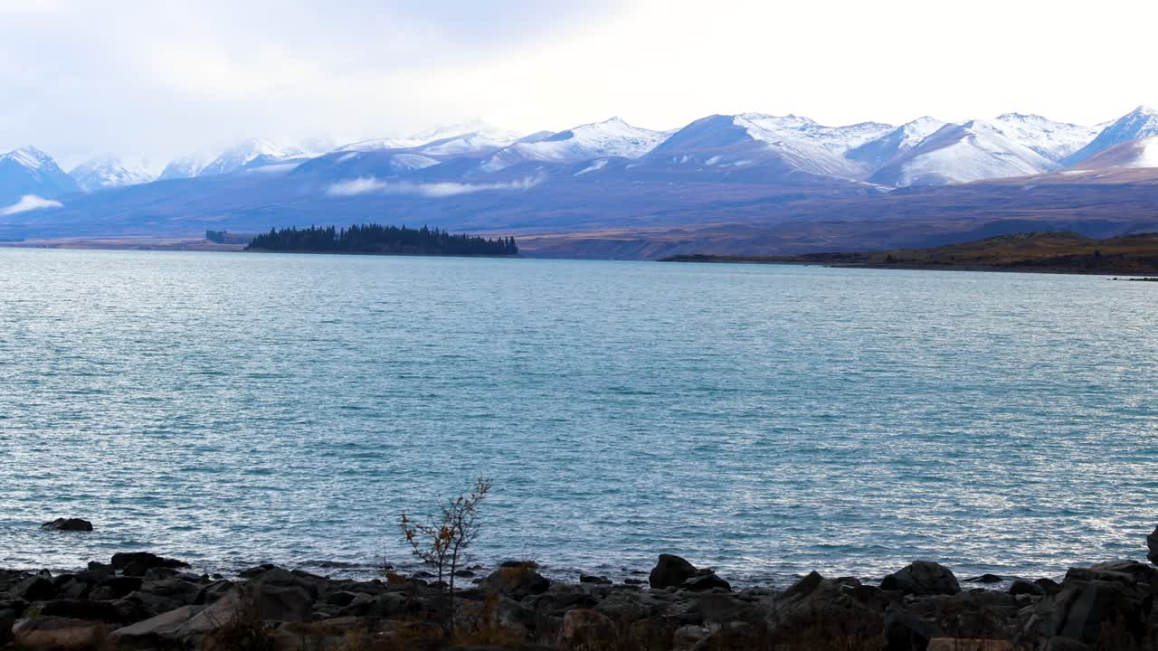 A tranquil view of Lake Tekapo with snow-capped mountains under soft, natural lighting. The scene captures calm water and distant hills