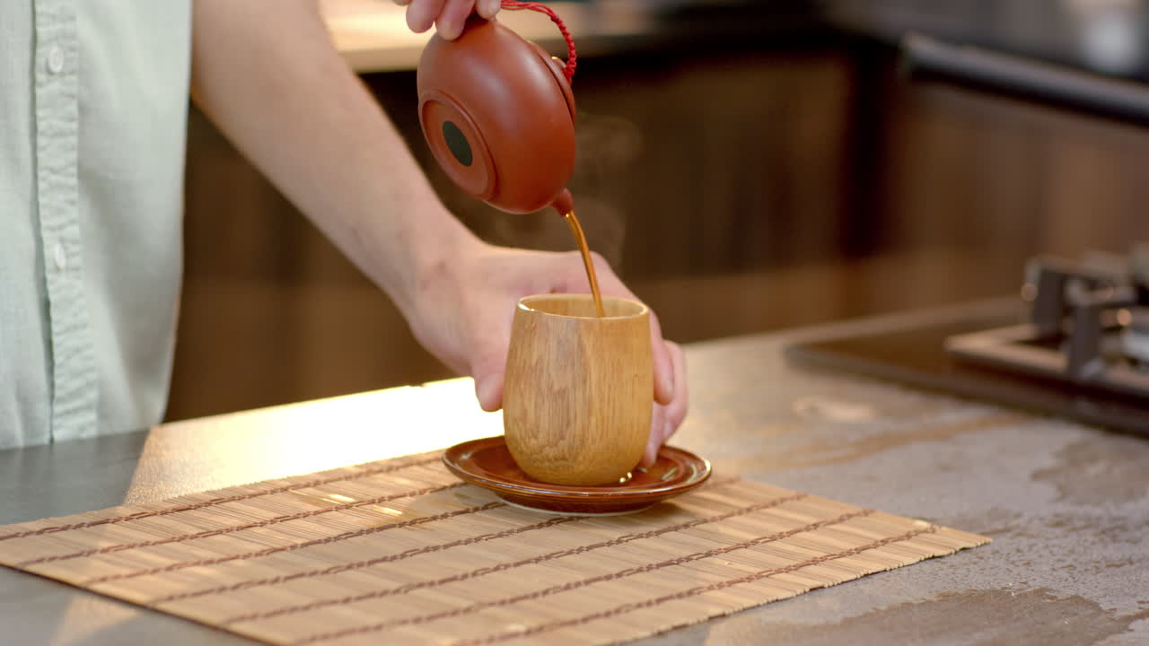 Pouring hot tea into wooden cup on bamboo mat in cozy kitchen