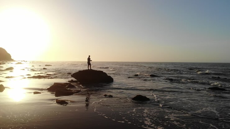 Um homem em silhueta ao nascer do sol observando as ondas do oceano quebrando na praia, na costa de Santa Bárbara, Califórnia. DRONE AÉREO.