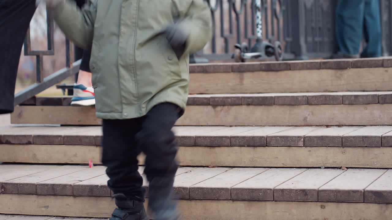 Child in winter coat runs down wooden park bridge stairs while woman bends to tie sneaker, scooter rests on upper step and other people stand nearby in calm outdoor setting with autumn background
