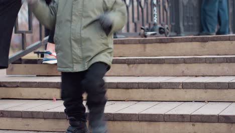child in winter coat runs down wooden park bridge stairs while woman bends to tie sneaker, scooter rests on upper step and other people stand nearby in calm outdoor setting with autumn background