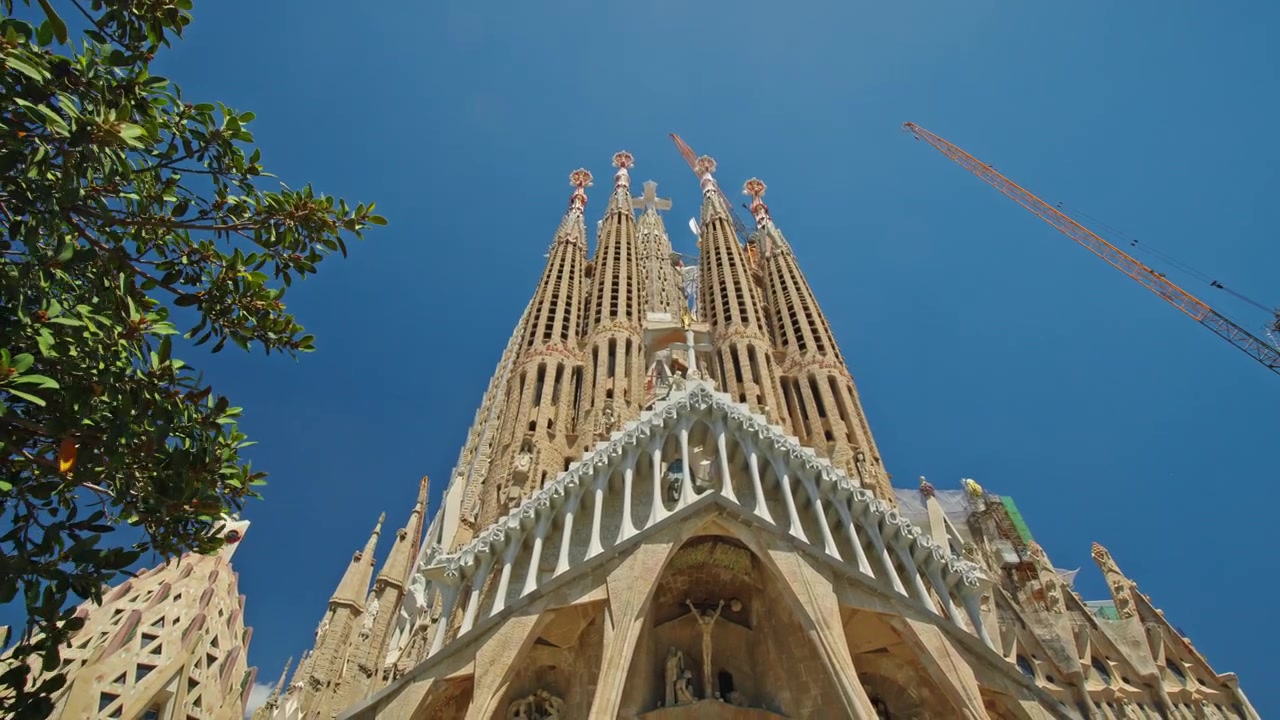 Vista architettonica dal basso della Basilica della Sagrada Familia a Barcellona, Spagna, capolavoro di Gaudí che si erge sopra il fogliame verde e una gru da costruzione contro un cielo azzurro limpido