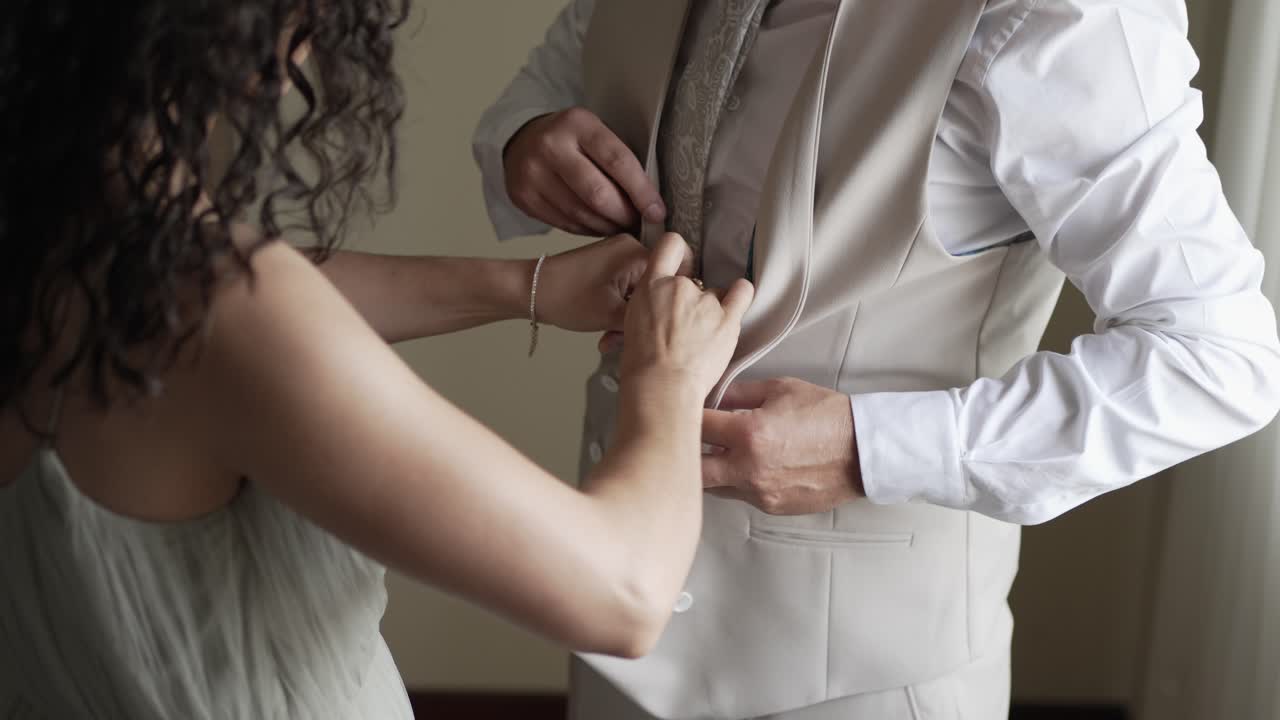 man in a beige vest is assisted by a woman with curly hair, both adjusting the buttons, suggesting a close, supportive moment during wedding preparations